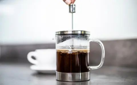 Eine Hand drückt den Stempel in eine French Press, um den perfekten Kaffee-Brew zuzubereiten. Eine Tasse wartet im Hintergrund.