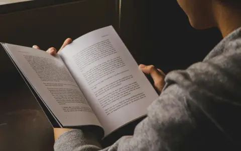 Osoba czyta książkę, skupiona na treści. W jej dłoniach spoczywa otwarta księga, a jej wzrok skierowany jest na strony, gdzie słowa tworzą historię, być może o trudnych wyborach i ich konsekwencjach, gdzie **inwersja** losu jest na porządku dziennym.