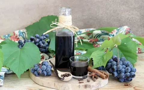 Mosto cotto ricetta: sciroppo scuro in bottiglia e bicchierino, con uva, spezie e cucchiaio di semi.