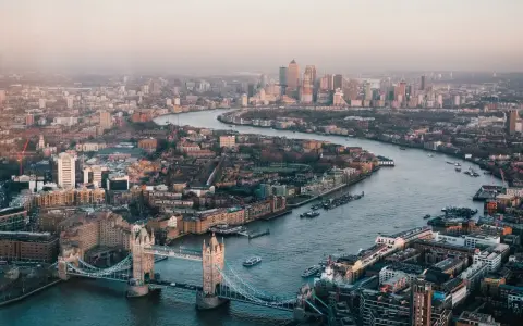 Stunning drone photography and videography captures London's iconic Tower Bridge and the winding Thames River, with the city skyline stretching into the hazy distance.