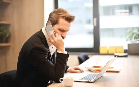 Mężczyzna w garniturze prowadzi ważną rozmowę przez telefon, siedząc przy biurku z laptopem i filiżanką kawy.