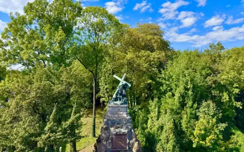 Pomnik na Jasnej Górze w Częstochowie, otoczony bujną zielenią drzew, pod błękitnym niebem z chmurami.