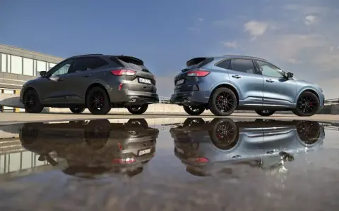 Dwa Fordy Kuga, jeden szary, drugi niebieski, stoją obok siebie na parkingu. Ich odbicia w kałuży tworzą surrealistyczny obraz. Oba modele to wersje hev.