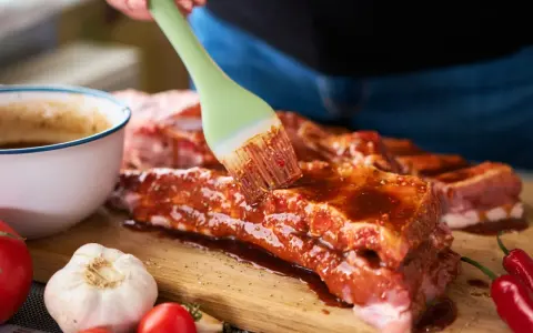 Pędzel rozprowadza aromatyczną marynatę do żeberek na surowym mięsie. Obok czosnek, pomidorki i papryczki chili.
