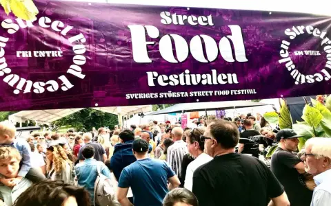 Stor folkmassa vid Sveriges största street food festival. En lila banderoll med texten "Street Food Festivalen" hänger över besökarna.