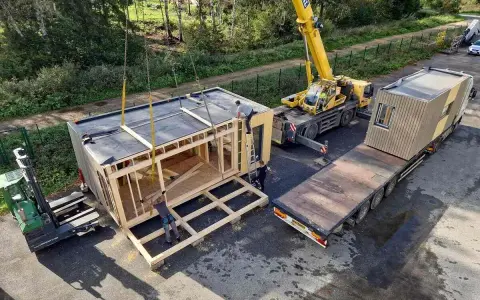 Une grue soulève un module de construction pour des maisons modulaires. Un chariot élévateur et un camion plateau sont également présents.