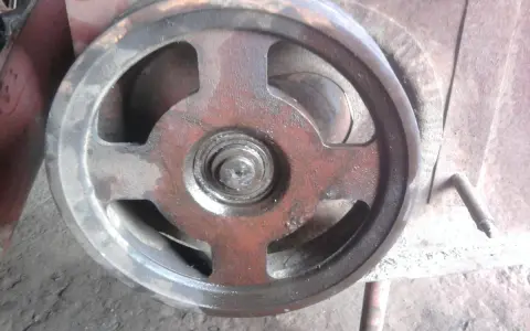 Metalowe koło pasowe kosiarki rotacyjnej, lekko zabrudzone i naoliwione, z widocznym łożyskiem pośrodku.