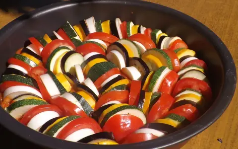 Un plat de légumes coupés en rondelles, prêt pour une recette de ratatouille digne du film. Tomates, courgettes, aubergines et oignons s'alternent.