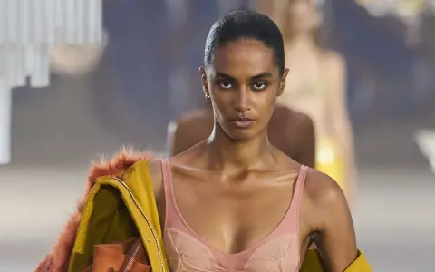 Modella in passerella con un audace reggiseno a vista color pesca, abbinato a una giacca gialla con pelliccia arancione.