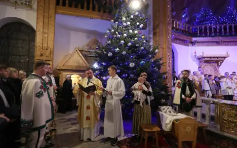 Pasterka w kościele w Zakopanem. Ksiądz czyta z księgi przy choince, obok ministranci i wierni w tradycyjnych strojach.