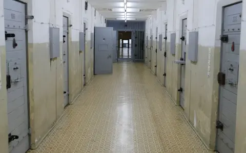 Korytarz więzienny z celami, gdzie może odbywać się tymczasowe aresztowanie. Widać drzwi z numerami, kraty i metalowe zabezpieczenia.