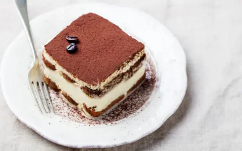 Una fetta di tiramisù, preparata con 500 g di mascarpone, guarnita con cacao e chicchi di caffè, su un piatto bianco con forchetta.