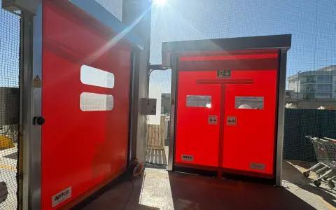 Due porte rosse automatiche, una aperta, mostrano come alzare una porta dal pavimento. Luce solare intensa.