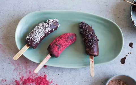 Tre hemmagjord glass bananer på pinne, doppade i choklad och toppade med kokos, kakaonibs och hallonpulver. Perfekt för barn!