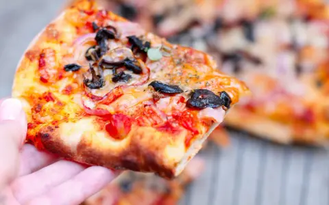 Kawałek pysznej pizzy funghi z pieczarkami, cebulą i pomidorami, trzymany w dłoni.