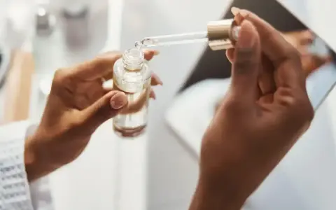 Dłonie trzymają buteleczkę z serum. Kropla z pipety, zawierająca kwas ferulowy, spada do buteleczki, przygotowując pielęgnację.