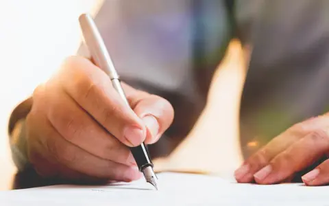 Dłoń pisze piórem po papierze, przygotowując dokument. Dowiedz się, jak napisać testament.