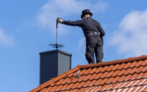 Kominiarz w czapce na dachu czyści komin. Zastanawiasz się, ile kosztuje przegląd kominiarski?
