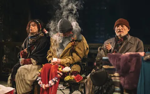 Antygona w Nowym Jorku: bohaterowie siedzą na ławce, otoczeni dymem i kwiatami, w ponurej atmosferze.