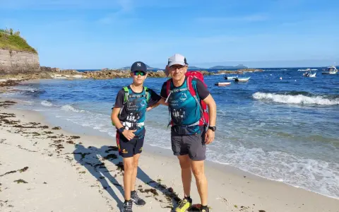 Dos excursionistas, padre e hijo, disfrutan de la playa en la etapa Baiona Vigo. El mar azul y el cielo despejado acompañan su aventura.
