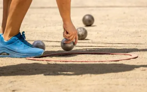 Une main saisit une boule de pétanque, prête à la lancer dans le cercle. Les règles du jeu de boules sont simples : viser le cochonnet.