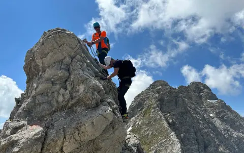 Zwei Bergsteiger meistern den **Hindelanger Klettersteig** auf felsigem Grat unter blauem Himmel mit Wolken.
