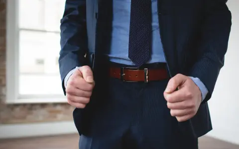 A man in a navy pinstripe suit and blue shirt wears a brown belt with black pants.