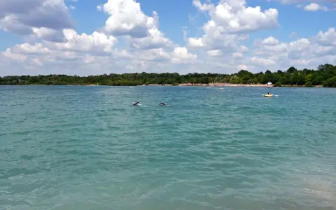 Schwimmer im türkisfarbenen Wasser des Beckum Sees, mit bewaldetem Ufer und Wolken am Himmel.