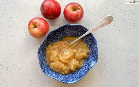 Compota de manzana casera en un cuenco azul, lista para disfrutar. Tres manzanas rojas frescas acompañan este delicioso postre.