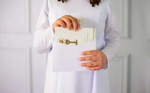 Dziecko w bieli wkłada kartkę z kielichem do koperty. To piękne krótkie życzenia na komunię, pełne symboliki.
