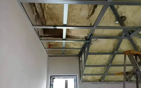 Konstrukcja podwieszanego sufitu z metalowymi profilami i pianką izolacyjną. Widać fragmenty ścian i okna.