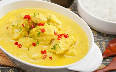 Delizioso pollo al curry con latte di cocco, guarnito con peperoncino rosso e erba cipollina. Accanto, una ciotola di riso bianco.