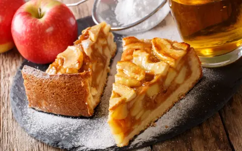 Deux parts de tarte aux pommes sur une ardoise, saupoudrées de sucre glace, avec des pommes et un verre de cidre.