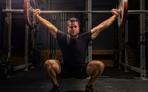 Ein Mann im tiefen Squat mit einer Langhantel über dem Kopf. Seine Muskeln sind angespannt, bereit für den nächsten Schritt im **power lifting**.