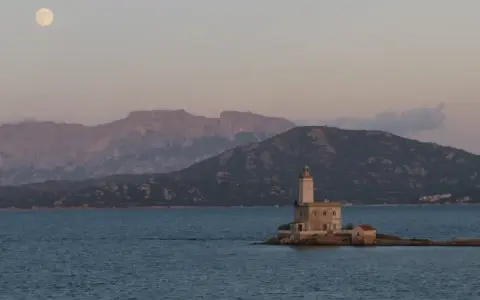 Faro solitario su un'isola sarda, con montagne sullo sfondo e luna piena. Perfetto per itinerari moto Sardegna 7 giorni.