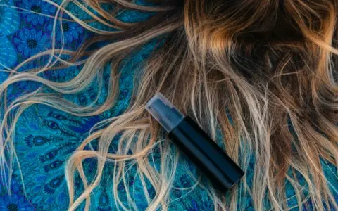 Czarny atomizer na tle błękitnego wzoru, obok rozpuszczonych, jasnych włosów. Idealny sposób, jak nawilżyć włosy.
