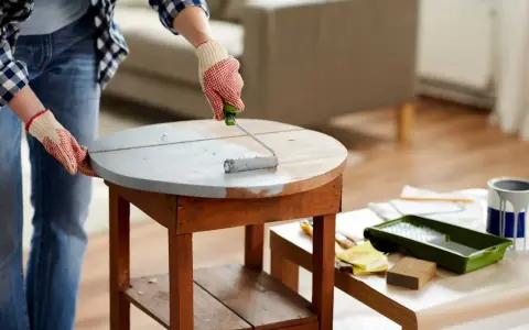 Une personne en jean et chemise à carreaux, portant des gants, utilise un rouleau pour appliquer une peinture grise sur une table ronde en bois. Le **décapage bois** est en cours pour un relooking.