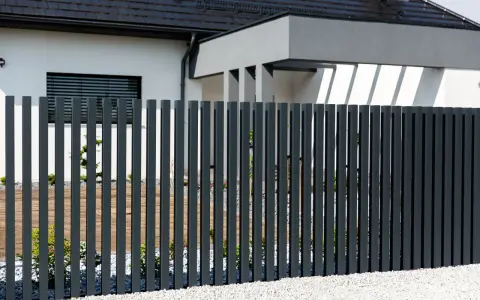 Nowoczesne, ciemnoszare ogrodzenie z pionowych profili to świetny pomysł na tanie ogrodzenie frontowe. Biały dom z czarnym dachem i żwirek dodają elegancji.