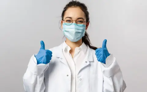 Uśmiechnięta pielęgniarka epidemiologiczna w masce i rękawiczkach pokazuje kciuki w górę.