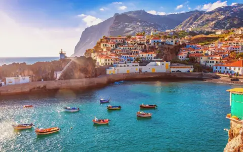 Bunte Häuser schmiegen sich an steile Hänge auf den Inseln bei Madeira. Kleine Boote schaukeln im azurblauen Wasser vor einem Leuchtturm.