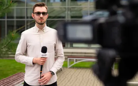Młody dziennikarz w okularach trzyma mikrofon z logo "FLESZ" przed kamerą.