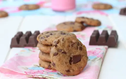 Una pila de galletas de chocolate caseras, con trozos de chocolate visibles, junto a barras de chocolate y un vaso de leche rosa.