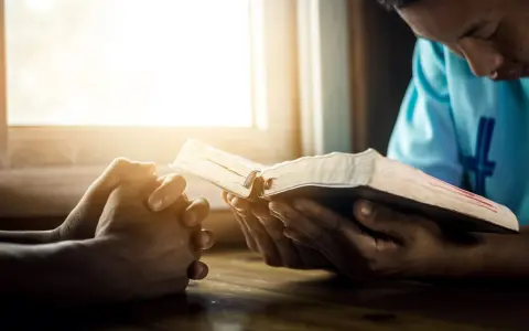Hands clasped in prayer beside an open Bible, bathed in warm light. A moment of reflection, perhaps contemplating prayer names.