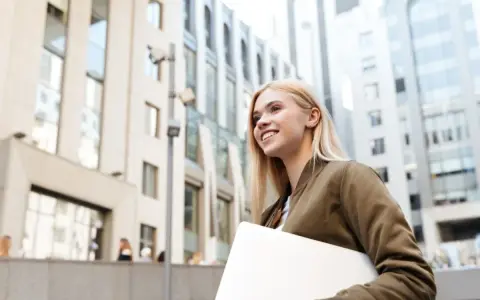 Giovane donna sorridente con laptop, cammina in una città moderna. Lavorare in Svizzera conviene, un futuro brillante ti aspetta.