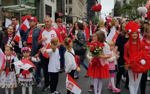 Dzieci i dorośli w strojach ludowych i z polskimi flagami maszerują ulicą. Widać serca z czerwonych balonów i kwiatów. Polacy w Szwecji świętują.