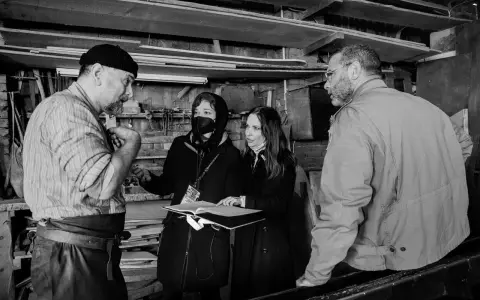 Foto di scena: un artigiano discute con due donne, una con mascherina, mentre un uomo osserva.