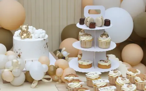 Biały tort z złotym misiem, babeczki i makaroniki na paterze, otoczone balonami w pastelowych kolorach.
