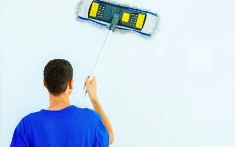 Hombre con camiseta azul limpiando paredes blancas con un plumero.