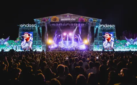 Gran concierto en el Jardín de las Delicias. Multitud vibrante ante el escenario iluminado, con pantallas gigantes mostrando al artista.