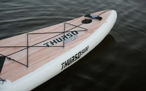 Ein aufblasbares SUP liegt im Wasser. Das Board hat eine Holzoptik und das Logo "THURSO SURF" ist gut sichtbar. Perfekt zum SUP lagern.